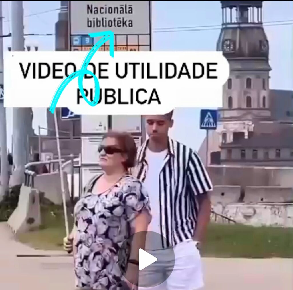 Cena de um vídeo onde se vê uma idosa cega no ponto de ônibus com uma bengala longa levantada. Ao lado, um rapaz a observa. Na imagem vemos a frase "Vídeo de Utilidade Pública". Uma seta azul direciona a atenção para um letreiro no ponto do ônibus onde lê-se " Nacionala biblioteka".