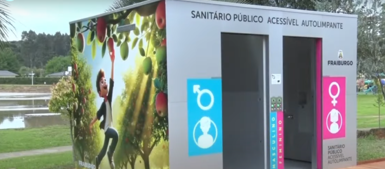 Imagem mostra uma cabine com 02 banheiros. Ela está instalada em um parque. Na cabine lemos: Sanitário público acessível autolimpante. Fraiburgo. Um dos lados da cabine é coberto com um adesivo que mostra o desenho de um garoto branco apanhando uma maçã. À frente, cada porta está indicada com adesivos azul e rosa e pictogramas específicos, referenciando o banheiro masculino e feminino, respectivamente.