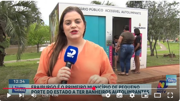 Imagem de uma reportatagem, onde a resporte aparece à frente de um banheiro acessível e autolimpante  