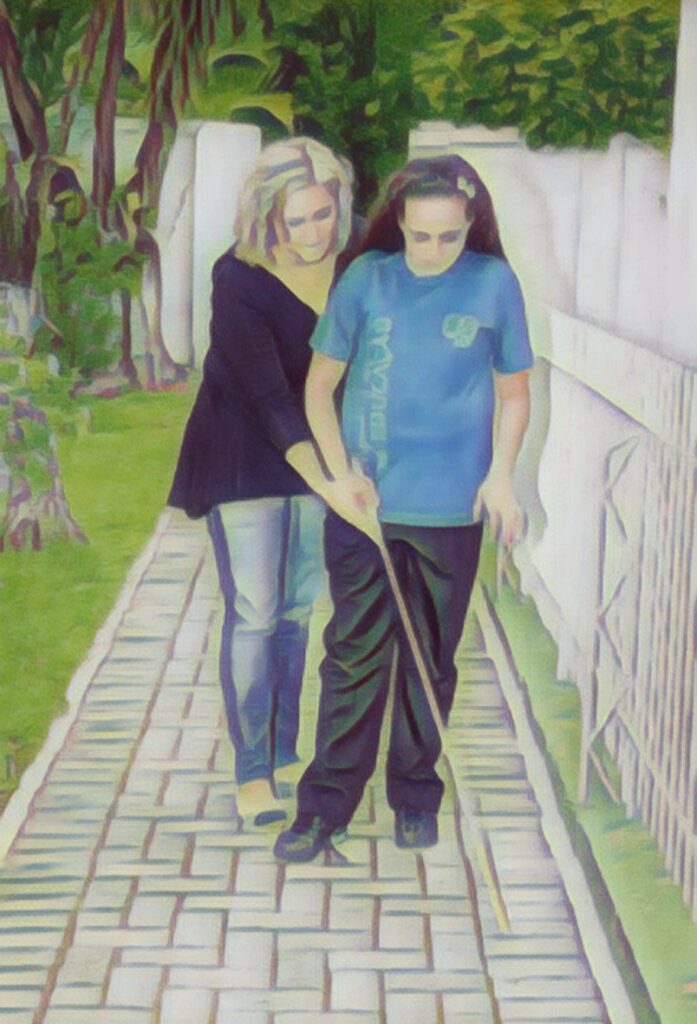 Imagem mostra professora de OM ensinando uma adolescente a andar com a bengala longa