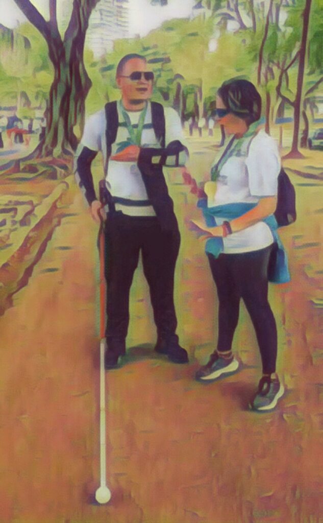 Imagem mostra uma professora de OM conversando com um rapaz que está segurando uma bengala longa. Eles estão em um parque