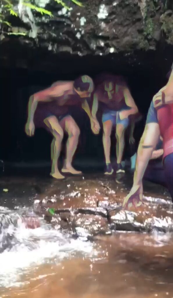 Dois homens abaixados estão saindo de uma espécie de caverna. Um deles segura a mão do outro e parece estar orientando-o. Eles caminham sobre um chão de pedras com água corrente vindo de uma cachoeira atrás deles.
