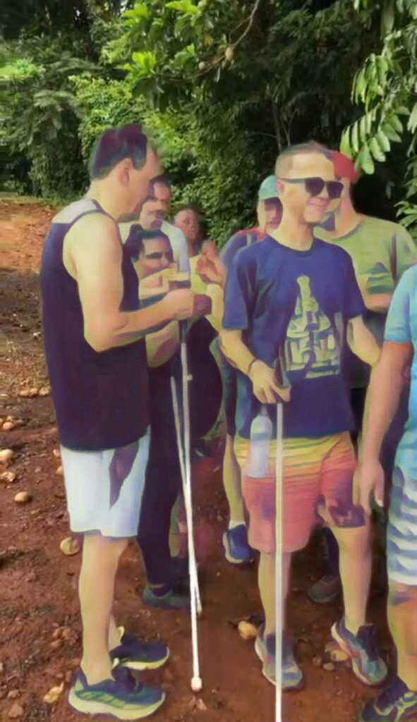 Um grupo de pessoas caminham em uma trilha na mata. Alguns estão usando bengalas longas.