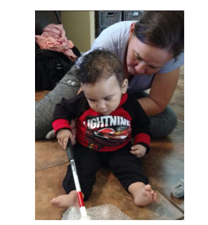 Imagem de uma mãe brincando com seu bebê. Ele está utilizando uma pequena bengala e passa a bengala sobre um plástico bolha.