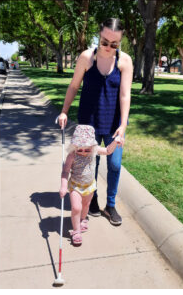 Imagem de uma mãe com sua bebê utilizando uma bengala longa. A bebê segura a bengala com uma mão e com a outra mão, segura a mão de sua mãe.