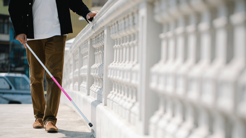 Imagem mostra uma pessoa com bengala longa andando. Com a mão livre, ela toca um muro branco.